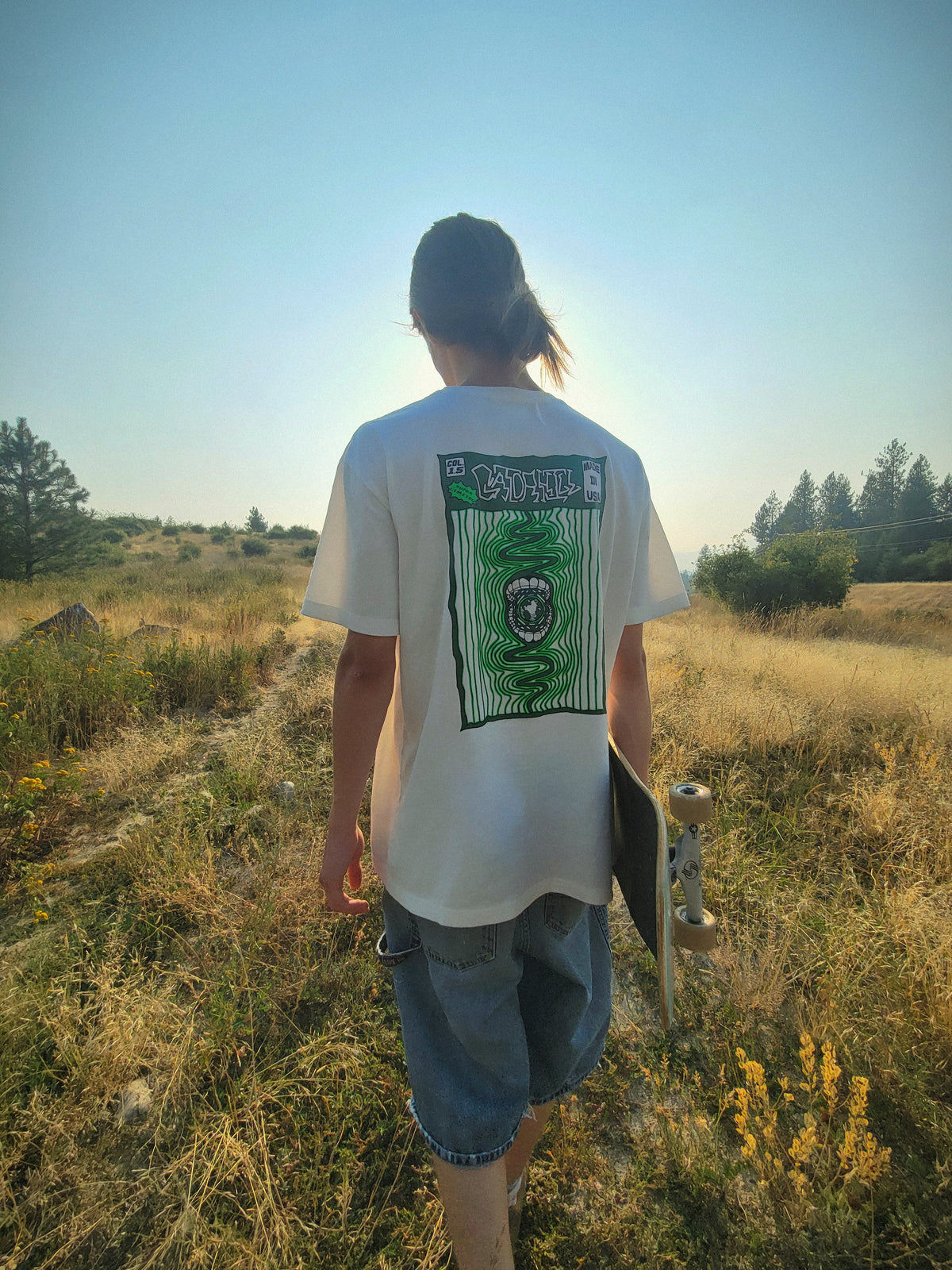 Person outdoors in the white Heavyweight Ripple Tee, back view showing the green gator ripple print, holding a skateboard